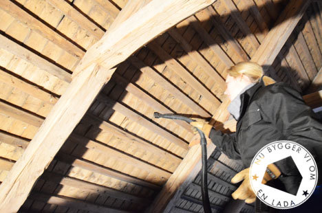 En person i arbeidsklær inspiserer treverket i taket på en lada. Sunlight treffer de solide trekonstruksjonene i taket.