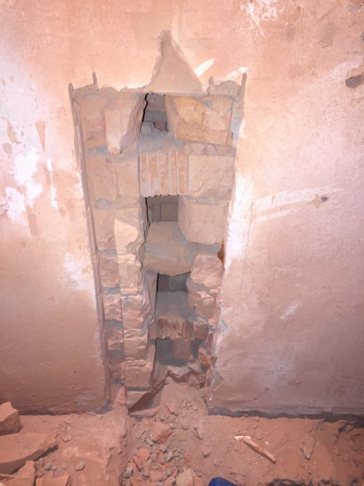 Bild av vegg med et stort hull hugget opp for å installere vann- og avløpsrør, med synlig luftspalte og murrester på gulvet.