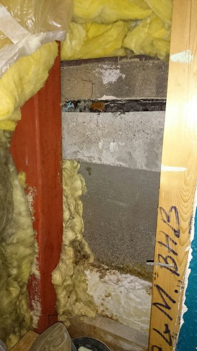 Eristetyn kellarin seinän sisällä näkyy betonilaatta ja punainen tukipalkki; eristevillaa ympärillä. Seinän päällä muita rakenteita.