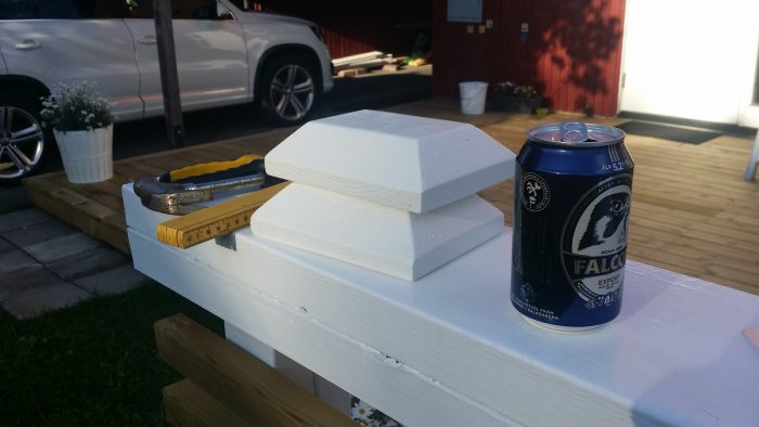 Tools and materials on a white wooden surface; a hammer, a folding ruler, two white wood caps, and a can of Falcon beer, with a car and deck in the background.