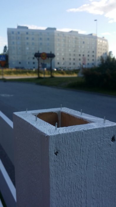 Close-up of a partially assembled rectangular wooden post with short nails sticking out, background shows a blurred building under blue sky.