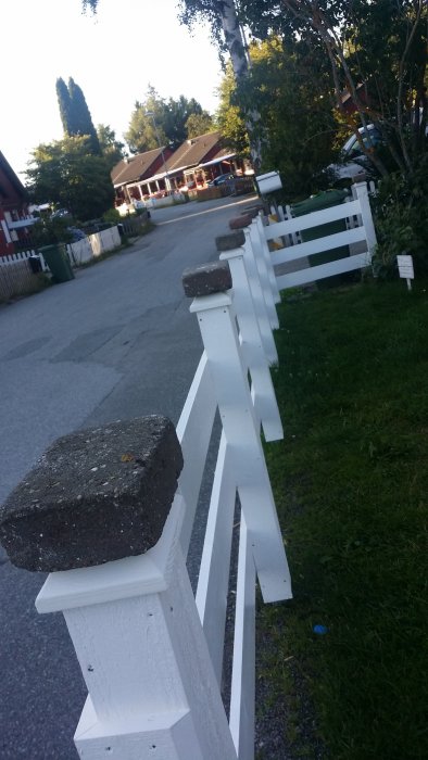 White fence with brick caps on posts, sunlight casts shadows; residential area visible in the background.