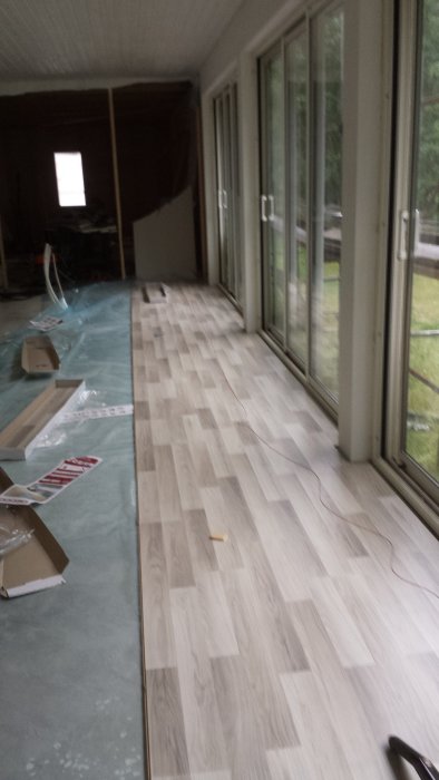 Wood laminate flooring with gap along large windows in a sunroom; a long underlay and scattered materials are visible, suggesting ongoing work.