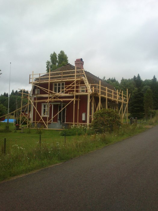 Red wooden house with DIY wooden scaffolding built around it for roof replacement; overcast sky in background.