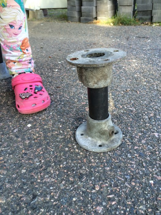 Rusty metal pipe connector on ground, ready to be cleaned and painted for reuse. A child's pink shoe and colorful pants visible beside.