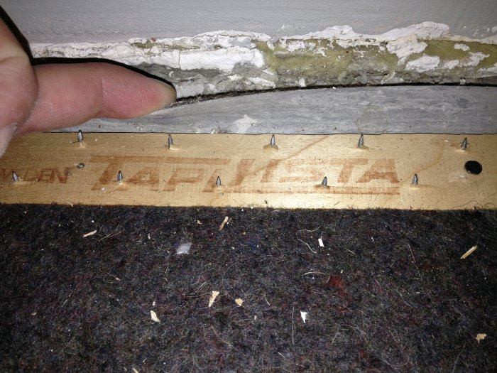 Fingeren løfter opp filttyg og plast som ligger over en betongoverflate. Små opp-ned spik er synlige langs kanten av treplaten.