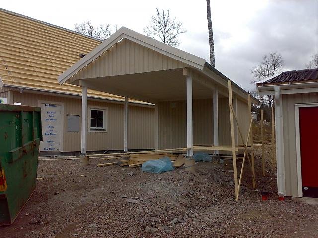En åpen carport under bygging med trestøtter, ved siden av et gult hus og en haug med grus og byggeavfall.