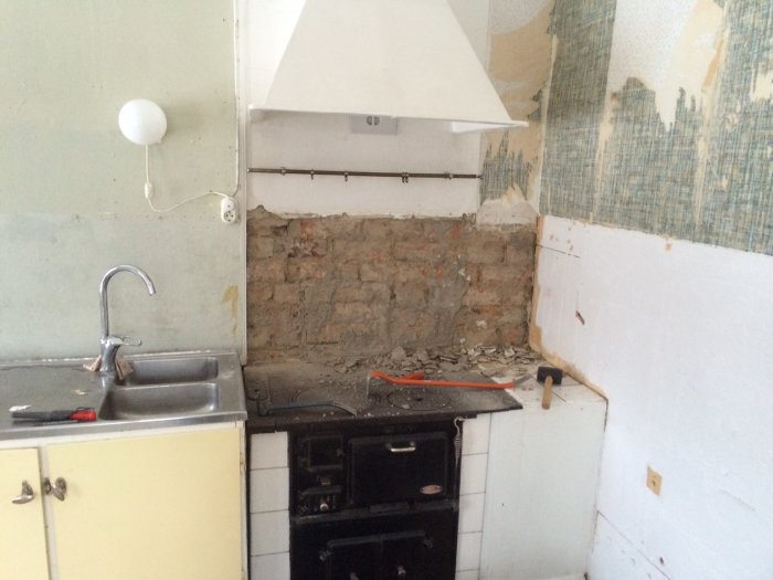 En kjøkkenvask og gammeldags svart komfyr med mursteinsvegg bak, klar til renovering etter å ha fjernet stygt flislagt område.