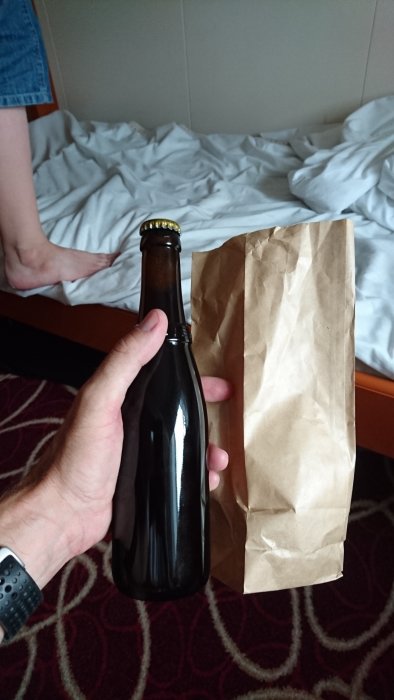 Person håller en flaska Westvleteren 12 öl bredvid en brun papperspåse, i ett slarvigt bäddat rum.