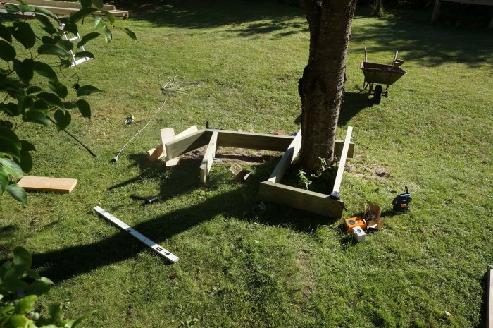 Trädgårdsbygge med vinkelbeslag monterade på träkonstruktion runt träd, verktyg och byggmaterial på gräs.