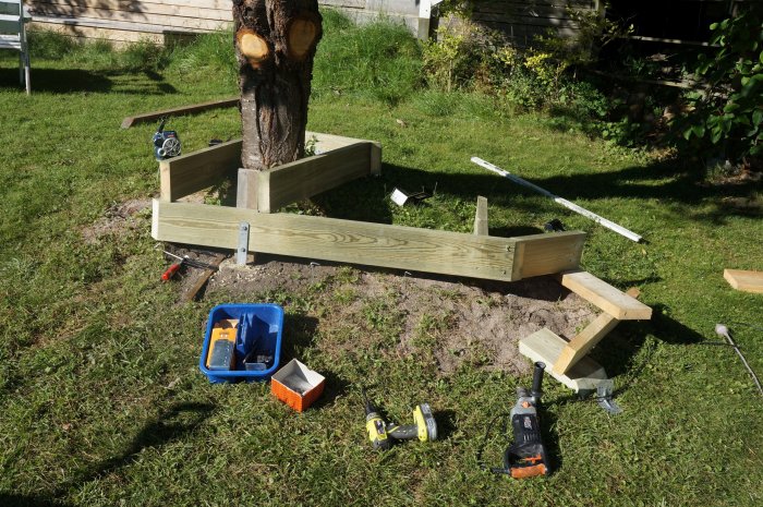 Trädgårdsbänk under konstruktion kring träd med verktyg och trämaterial på gräsmatta.