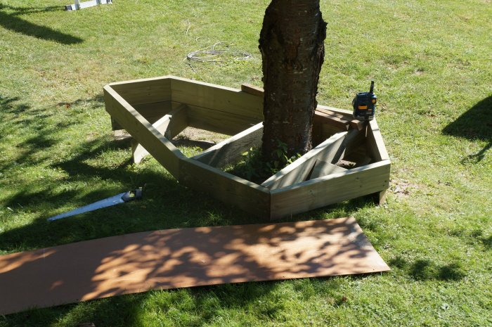 Trästomme av byggprojekt runt ett träd i en trädgård, masonitskiva och verktyg på marken.