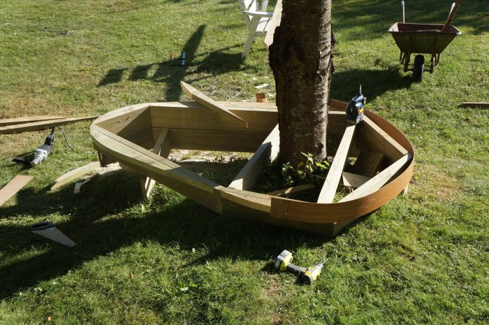 Njureformad träkonstruktion med masonitremsor under byggnation utomhus, verktyg och skottkärra i bakgrunden.