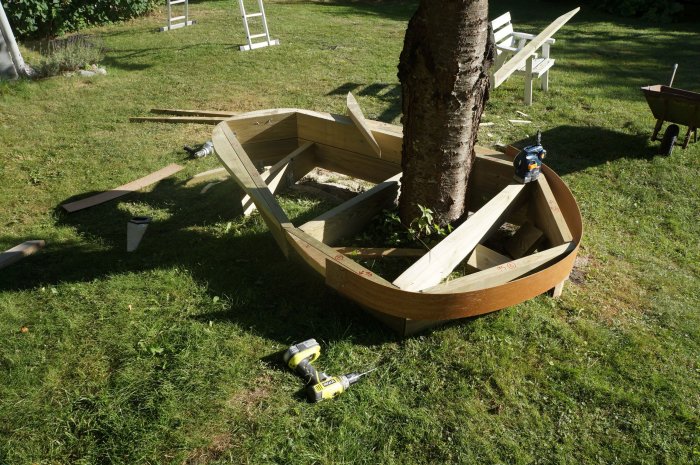 Byggprojekt med masonitremsor formade som en njure runt ett träd, halvfärdigt med verktyg på gräsmattan.