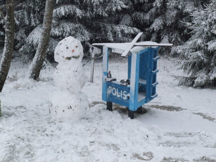 Ett snöfall, en snögubbe bredvid en blå träkiosk märkt "POLIS", i ett skogligt vinterlandskap.