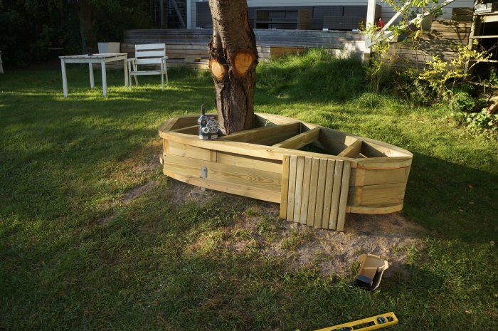 Halvfärdig träkonstruktion för inklädnad runt träd med 1 cm mellanrum till marken, trädgårdsverktyg syns.