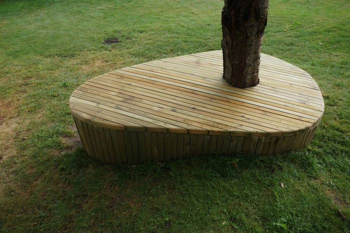 Bänkyta byggd av diagonalt liggande 45 x 45 mm brädor runt ett träd på en gräsmatta.