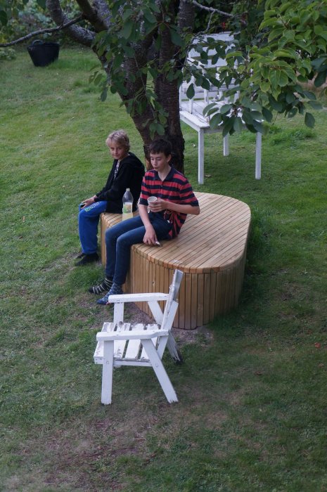 Två personer sitter på rund trädgårdsbänk och fikar i en trädgård.