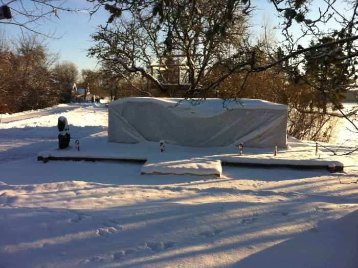 Pallar med Leca-block täckta av presenning på en snötäckt mark i vintersol.