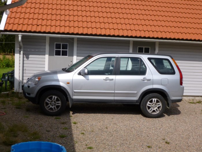 En äldre silverfärgad SUV parkerad framför ett vitt hus med röd takpannor.