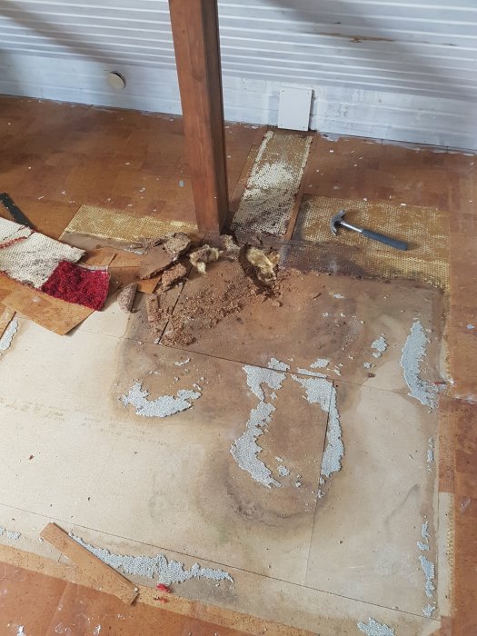 Golvspånskivor med fuktskador och smulande delar nära en träpelare.