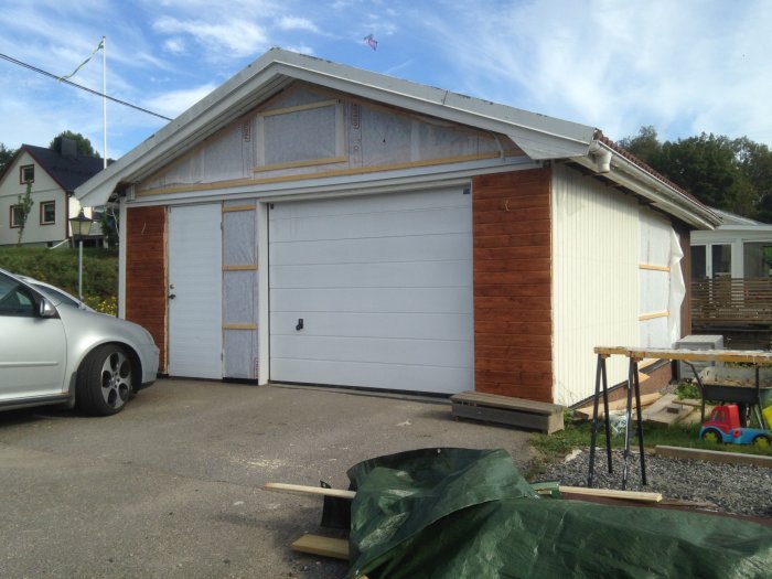 Garage med halvfärdig panelvägg i trä, delvis isolerad, bredvid en vit garageport och en parkerad bil.