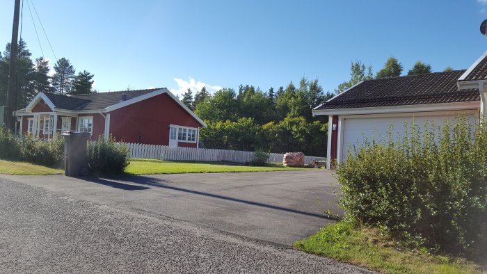 Rödmålad villa med tillhörande garage och vit staket, plan gräsmatta och buskar, klar blå himmel på dagen.