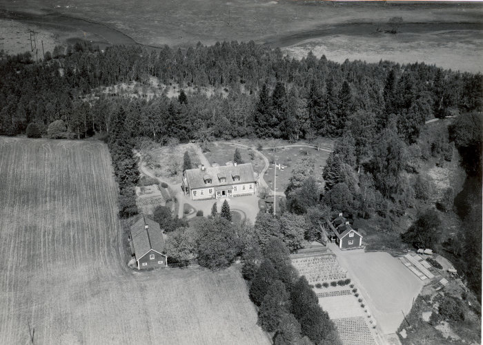 Svartvit flygbild över lantegendom omgiven av åkrar, skog och trädgårdsodlingar. Vintage estetik.