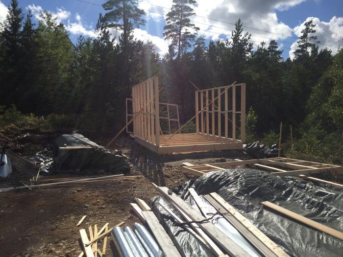 Delvis byggd trähustomme utomhus med takbjälkar på plats, omgiven av skog och byggmaterial på marken.