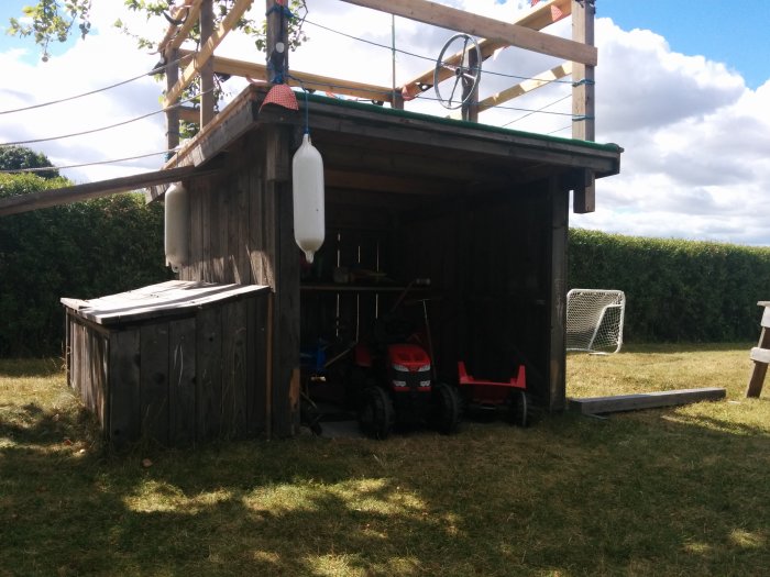 En ombyggd carport för barn med leksaker, hyllor, klätterribbor och konstgräsmatta på taket.