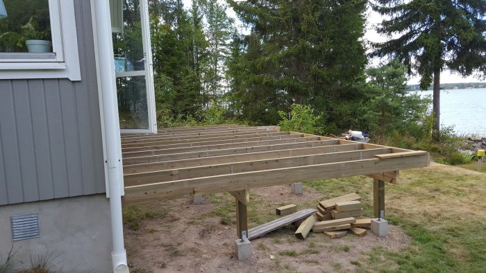 Pågående bygge av en 7x3.6m stor altan utanför ett hus med utsikt över en sjö, vissa reglar saknas.