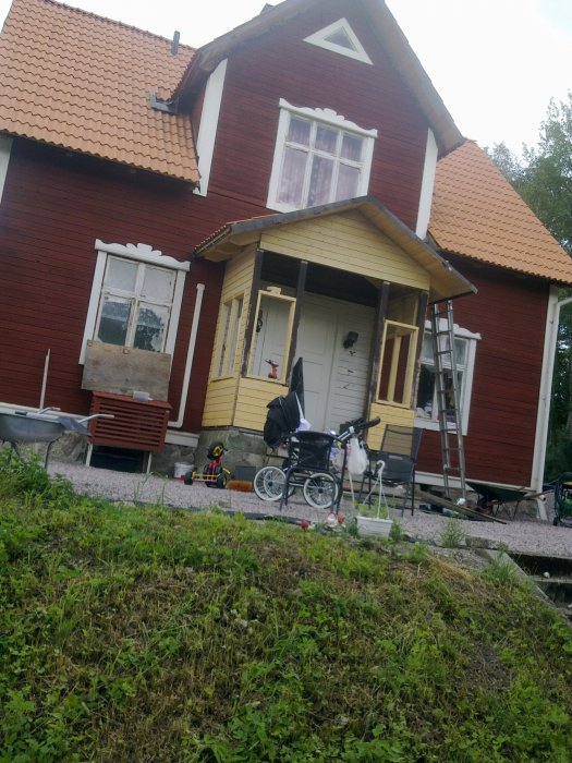 Röd trävilla med en farstu under konstruktion, leksaker och en barnvagn framför huset.