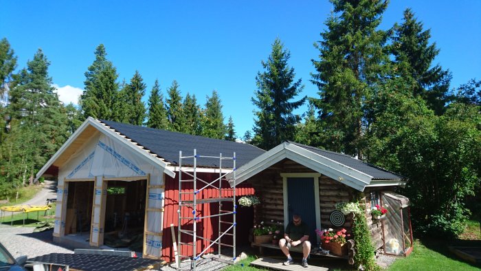 Två garage vid en stuga under renovering, ena med ny takplåt och byggställningar, den andra med sittande person.