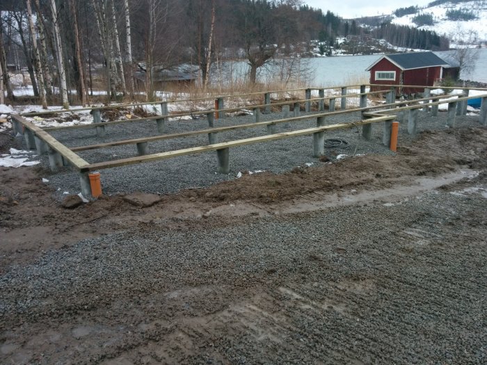 Grund till hus med stolpar och bjälkar på grus, snötäckt mark och röd stuga vid vattnet i bakgrunden.