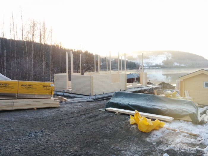 Byggplats med husväggar och byggmaterial i förgrund, sjö och snöklädda kullar i bakgrund.