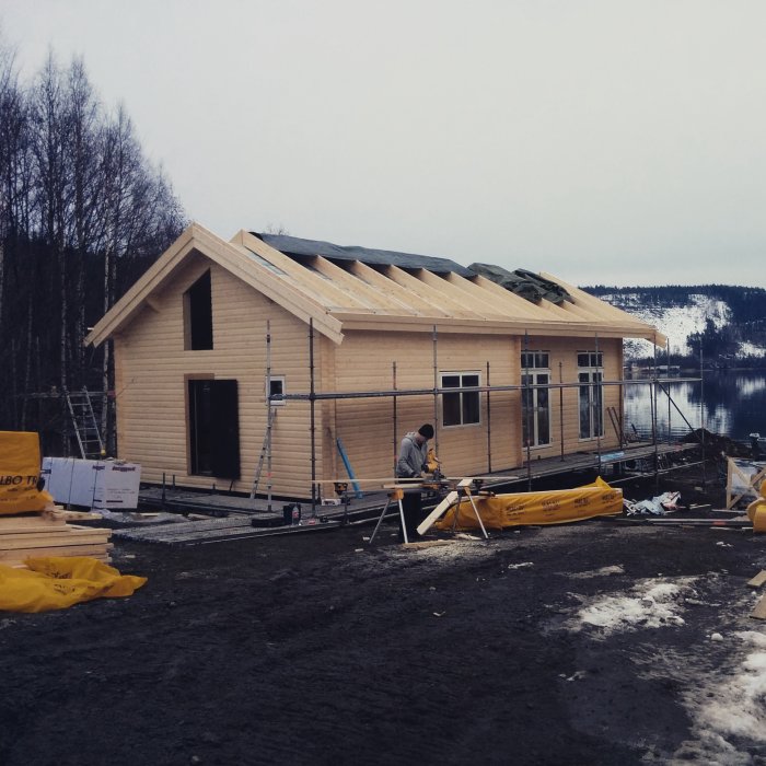 Ett halvfärdigt trähus under konstruktion vid vattnet med synligt ställningsarbete och byggmaterial runtomkring.