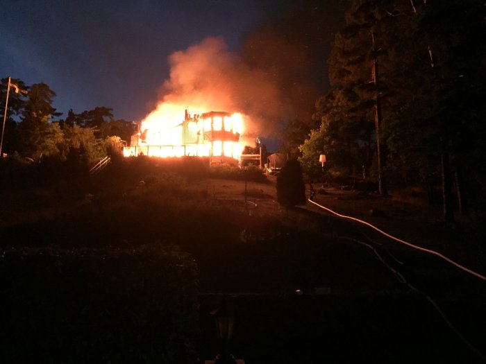 Brand på nattetid förstör hus, med flammande eld och rök, intakt terrass syns i förgrunden.