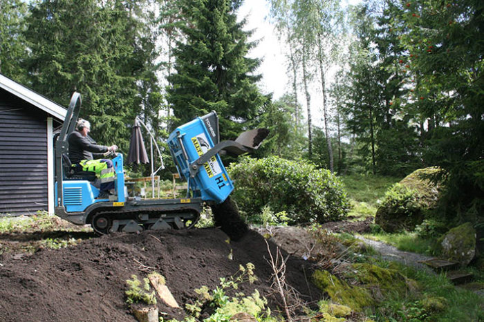 Person använder en kompaktlastare för markarbete i en trädgård med grönska och träd i bakgrunden.