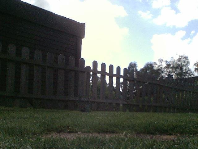 Uppdaterad gard med kamera, visar lågt perspektiv av trästaket och gräs med suddig bakgrund.