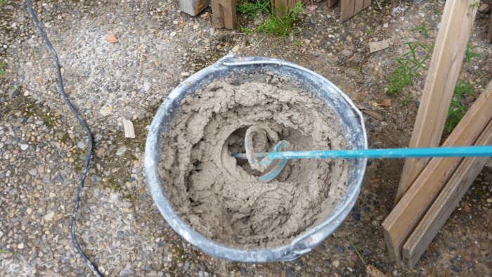 Hink med blandad cement och vatten med en blå blandare i, på en grusig yta utomhus.