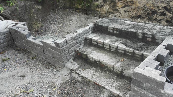 Halvfärdig stödmur under konstruktion med mörkgrå betongstenar och synliga ojämnheter.