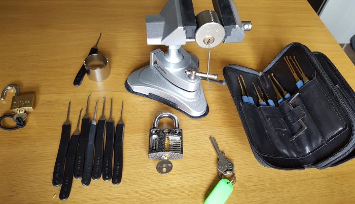 Lock picking kit with tools and practice locks on a table, including a transparent padlock and a vice holding a cylinder lock.