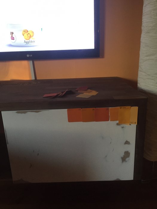Hembyggd TV-möbel med vita luckor och färgprover, framför en TV på väggen.
