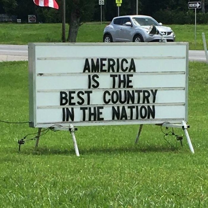 Skylt med texten "AMERICA IS THE BEST COUNTRY IN THE NATION" vid en gräsmatta nära väg med bil i bakgrunden.