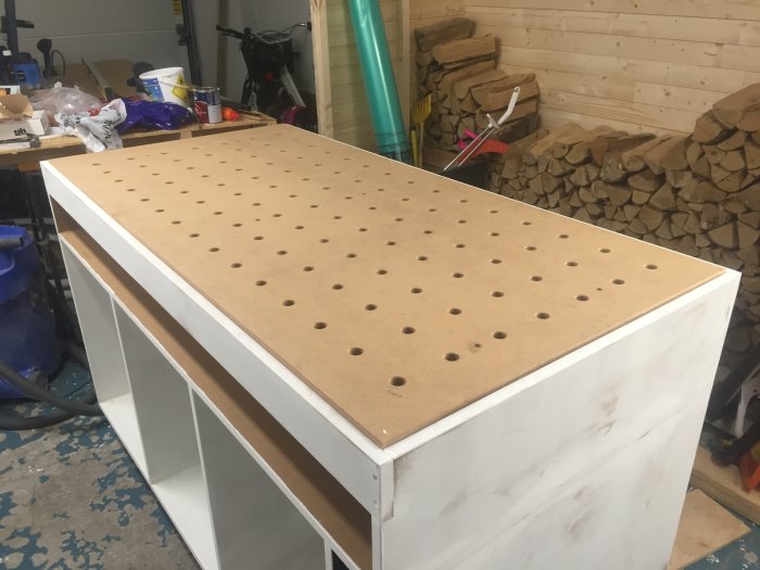 Verktygsbänk med perforerad skiva monterad på fyrkantiga benstöd i ett garage med vedstapel i bakgrunden.