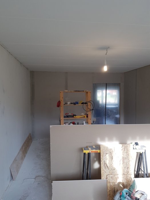 Ett oinrett rum med synliga gipsskivor och spacklade skarvar, byggmaterial och en arbetsbänk syns.