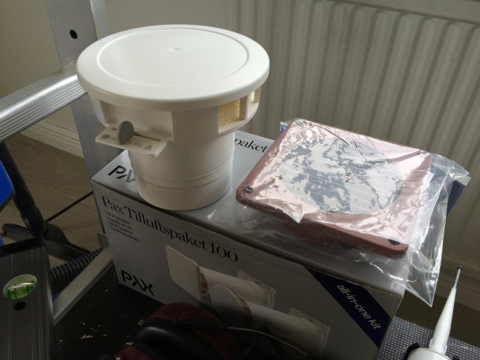 Pax tilluftspaket och falurött galler för ventilation ännu i förpackningen på ett arbetsbord.
