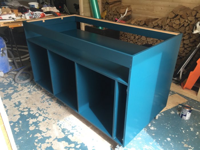 Nymålad blå köksö snickeri i ett verkstadsrum med verktyg och vedstapel i bakgrunden.