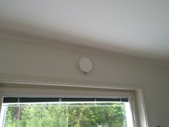 Ventilationshål med förlängare ovanför ett fönster i en vitmålad vägg, i ett rum med skogsbakgrund.