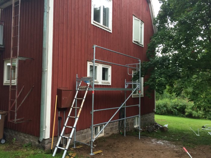 Nyuppsatt byggställning med justerbara ben och sidoräcken vid röd husfasad, flankerad av en stege och byggmaterial.